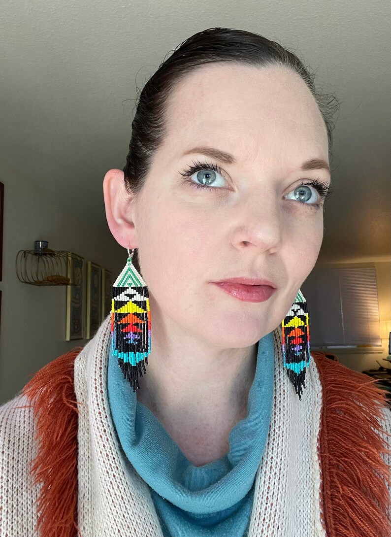 May include: A woman wearing a blue turtleneck sweater and a white and orange cardigan is wearing a pair of long, colorful beaded earrings. The earrings are black, white, green, blue, orange, yellow, and red.