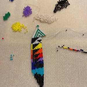 May include: A colorful beaded feather earring with a black, white, green, blue, yellow, orange, and red pattern. The earring is partially strung on a white thread with beads of various colors scattered around it.