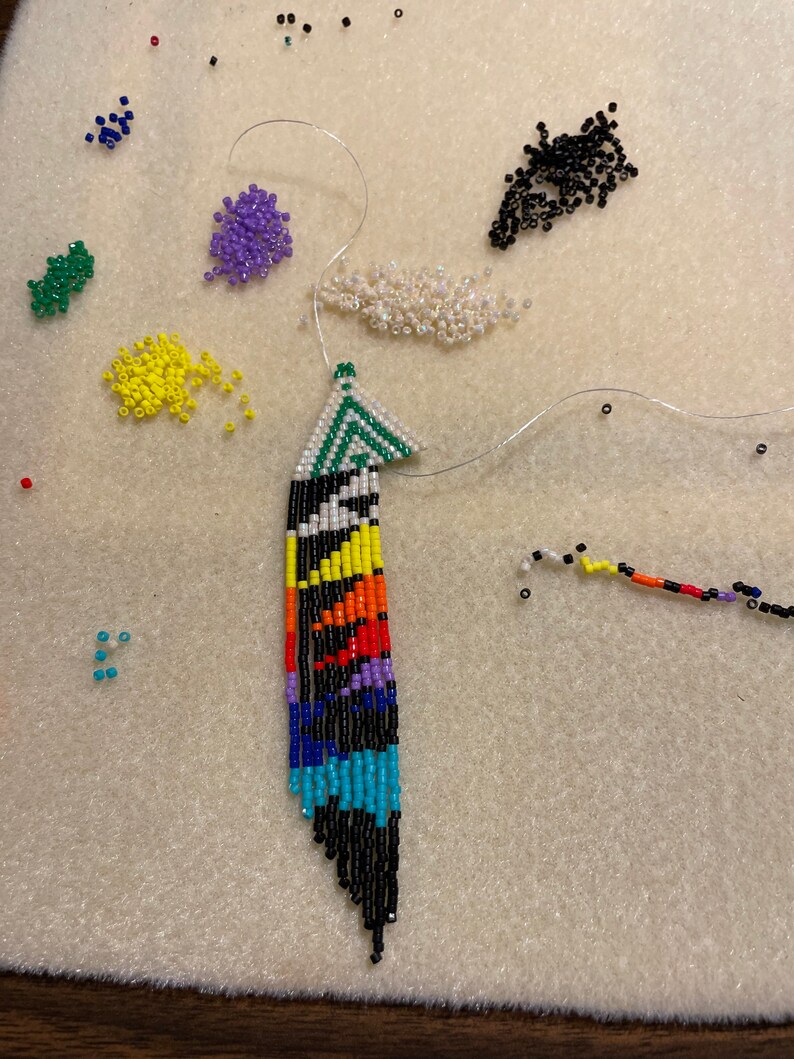 May include: A colorful beaded feather earring with a black, white, green, blue, yellow, orange, and red pattern. The earring is partially strung on a white thread with beads of various colors scattered around it.