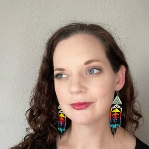 May include: A woman wearing a black shirt with long, curly brown hair. She is wearing a pair of long, colorful beaded earrings with a geometric pattern.