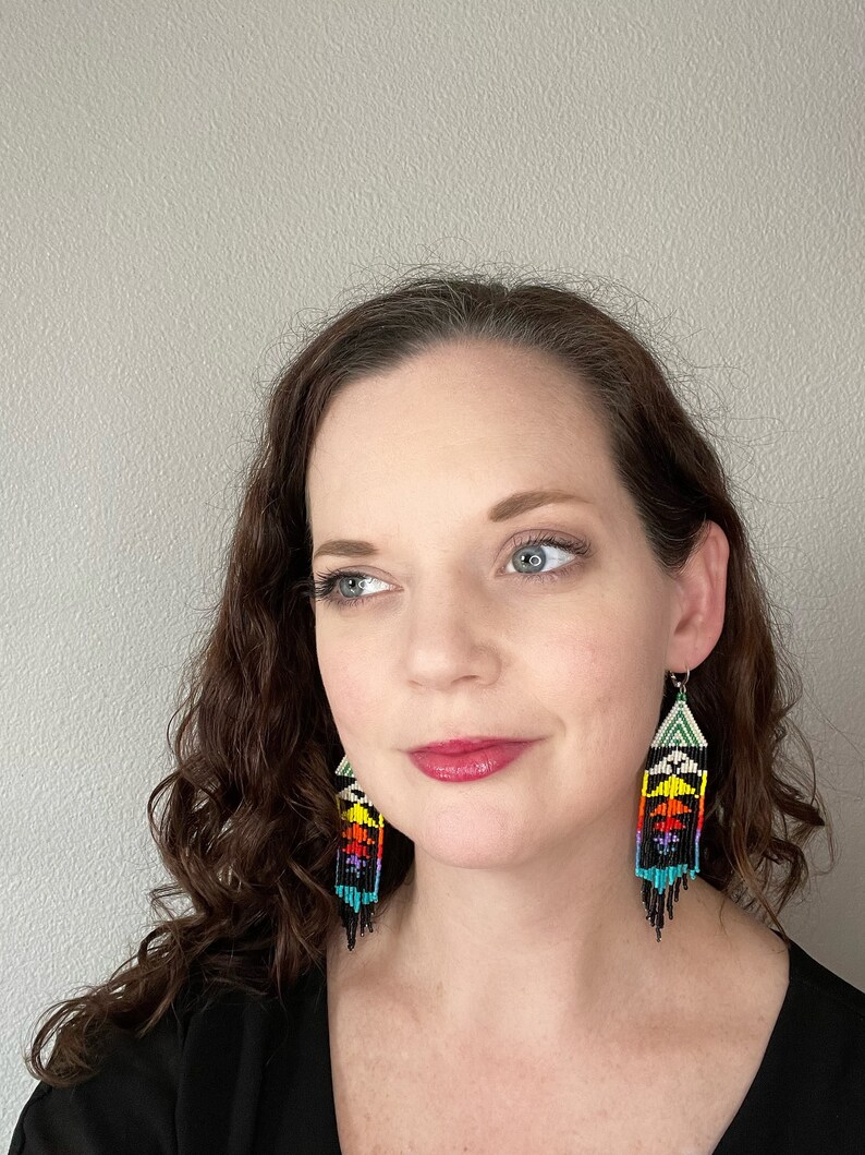 May include: A woman wearing a black shirt with long, curly brown hair. She is wearing a pair of long, colorful beaded earrings with a geometric pattern.