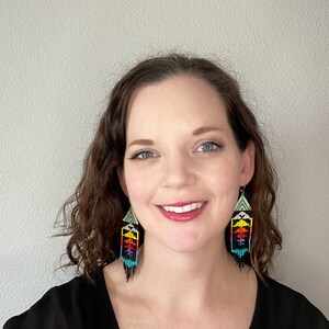 May include: A woman with curly brown hair wearing a black top and colorful beaded earrings. The earrings are long and dangle, with a geometric pattern in shades of red, yellow, green, blue, and black.