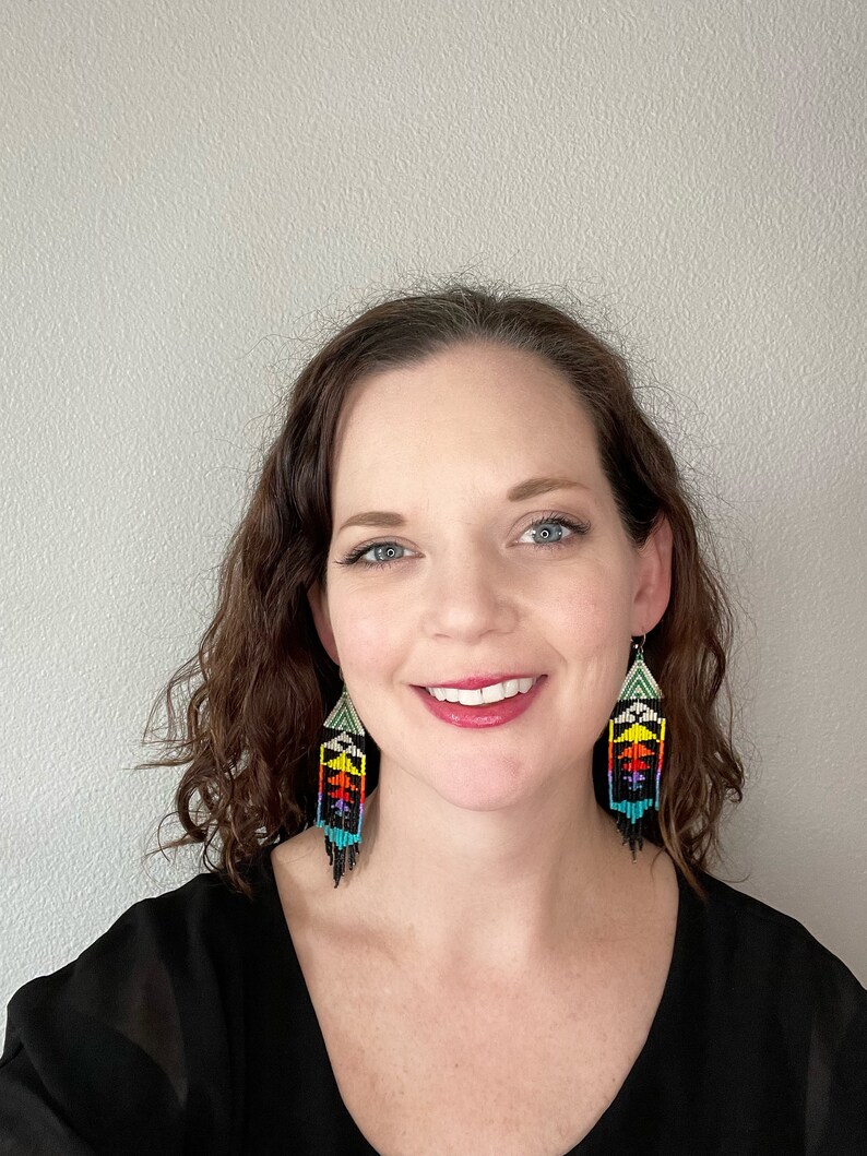 May include: A woman with curly brown hair wearing a black top and colorful beaded earrings. The earrings are long and dangle, with a geometric pattern in shades of red, yellow, green, blue, and black.