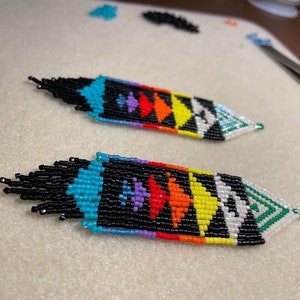 Pode incluir: Um par de brincos de contas com um padrão geométrico em preto, branco, turquesa, verde, amarelo, laranja, vermelho e roxo. Os brincos são longos e pendentes.