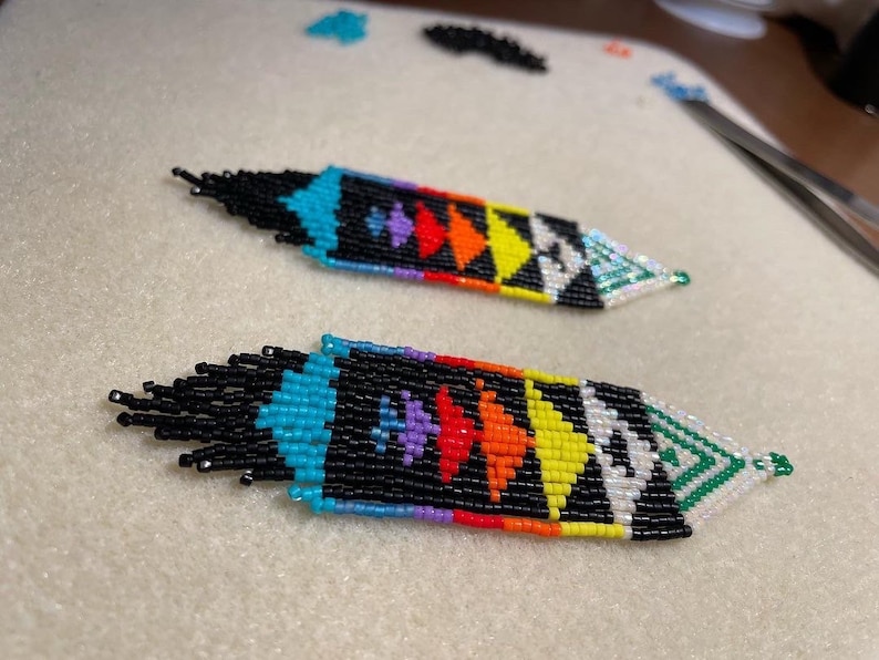 May include: A pair of beaded earrings with a geometric pattern in black, white, turquoise, green, yellow, orange, red, and purple. The earrings are long and dangle.