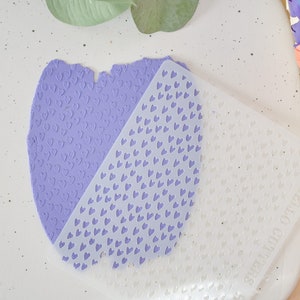 May include: A white plastic heart-shaped stencil is placed on top of a purple heart-shaped piece of clay. The clay has been embossed with a heart pattern using the stencil.