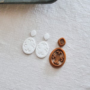 May include: Two white oval-shaped earrings with a leaf design and a brown plastic mold of the earrings with a leaf design.