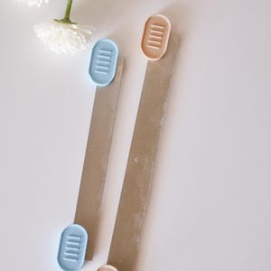 May include: Two sets of craft supplies are displayed on a white surface. Each set includes a rectangular piece of cardboard and an oval-shaped, ridged plastic piece. One set has a light blue plastic piece, and the other has a peach-colored plastic piece. White flowers are in the background.