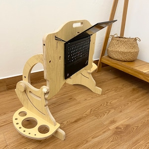 May include: A light-colored wooden laptop stand with an adjustable vertical panel and a base featuring circular cutouts. A black laptop is mounted to the center of the vertical panel using black elastic cords, allowing for an elevated viewing angle.