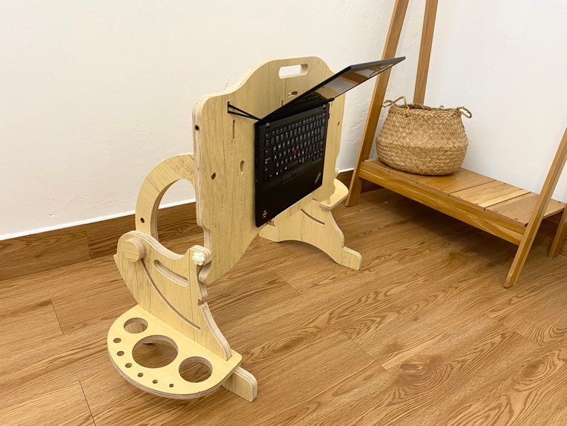 May include: A light-colored wooden laptop stand with an adjustable vertical panel and a base featuring circular cutouts. A black laptop is mounted to the center of the vertical panel using black elastic cords, allowing for an elevated viewing angle.