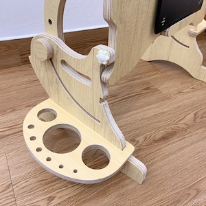 May include: A light-colored plywood laptop stand featuring an adjustable frame with white plastic knobs and a semi-circular base attachment with various circular cutouts of different sizes for holding accessories or writing instruments.