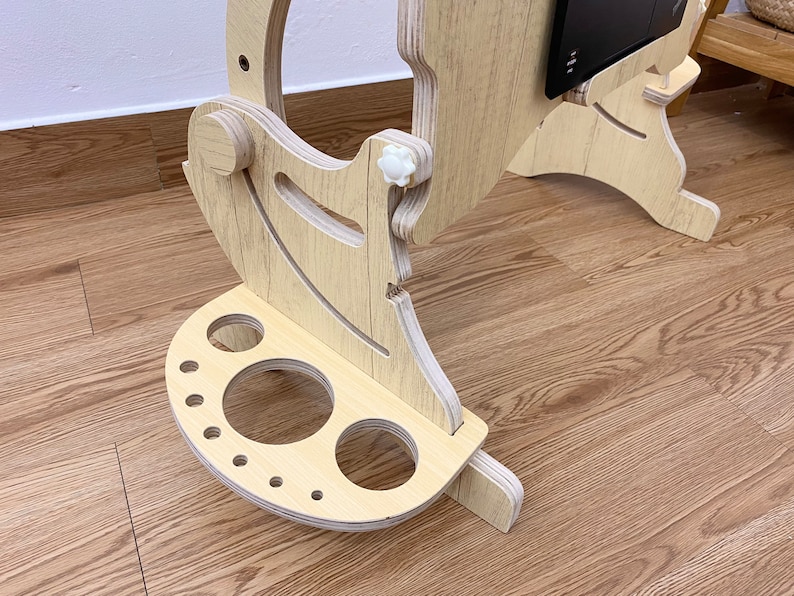 May include: A light-colored plywood laptop stand featuring an adjustable frame with white plastic knobs and a semi-circular base attachment with various circular cutouts of different sizes for holding accessories or writing instruments.