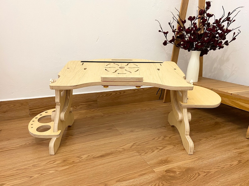 May include: A light-colored wooden laptop desk with an adjustable surface, featuring a central circular cutout design, a small front ledge, and two side attachments, one with circular holes and one with a smooth, rounded surface extension.