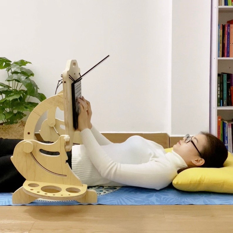 May include: A wooden laptop stand with a person lying on a blue mat using the stand. The stand has a curved design and a shelf for a laptop. The person is wearing a white sweater and has glasses on.