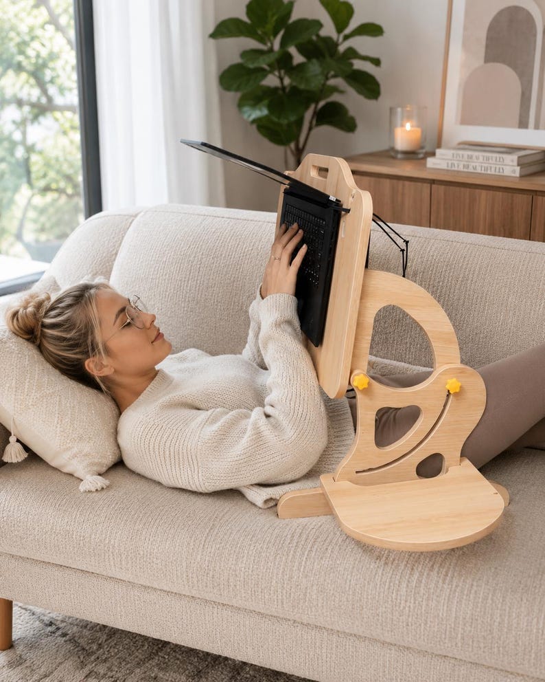 May include: A light-colored wooden laptop stand with an adjustable frame, featuring yellow knobs for height and angle customization. The stand includes a flat base, a vertical support structure with cutouts, and a platform to hold a black laptop.