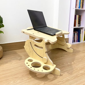 May include: A wooden laptop stand with a built-in cup holder and a shelf for a mouse. The stand is light brown and has a natural wood finish. A black laptop is on the stand.