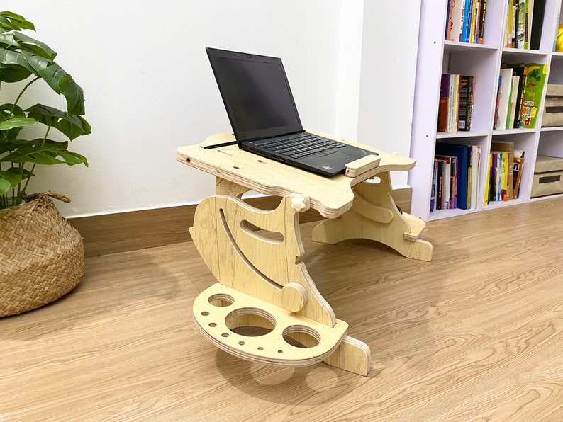 May include: A wooden laptop stand with a built-in cup holder and a shelf for a mouse. The stand is light brown and has a natural wood finish. A black laptop is on the stand.
