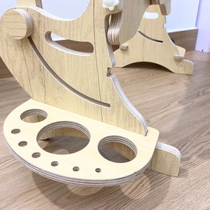 May include: A wooden stand with a curved top and a base with multiple circular cutouts. The stand is made of light wood with a wood grain pattern.