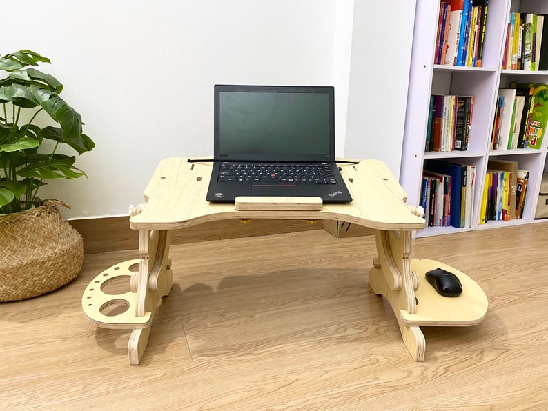 May include: A wooden laptop stand with a built-in mouse pad and cup holders. The stand is light brown and has a laptop on top of it.