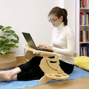 May include: A person sits on a blue yoga mat using a wooden laptop stand with a cup holder. The stand is designed to be used while sitting on the floor. The person is wearing a white turtleneck sweater and glasses.