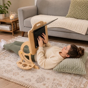 May include: A light-colored wooden laptop stand with an adjustable angled support, a handle cutout at the top, and a base featuring multiple circular cutouts for storage. The stand holds a black laptop in an upright position for use while lying down.