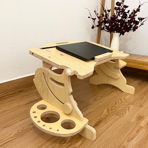 May include: A light-colored wooden laptop desk with an adjustable surface and a black laptop placed on top. The desk features a side attachment with circular cutouts for holding items and a curved support structure with visible adjustment knobs.