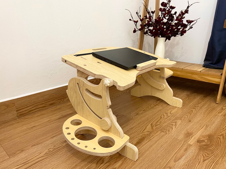 May include: A light-colored wooden laptop desk with an adjustable surface and a black laptop placed on top. The desk features a side attachment with circular cutouts for holding items and a curved support structure with visible adjustment knobs.