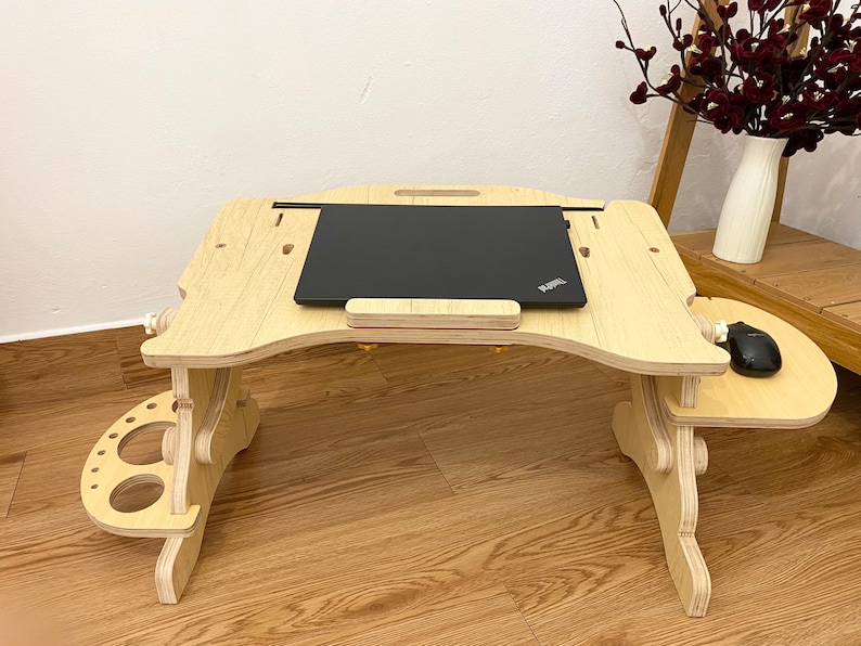 May include: A light-colored wooden laptop desk with an adjustable surface, a black laptop, and a black computer mouse. The desk features a side platform for a mouse and a secondary side attachment with circular cutouts for holding various items.