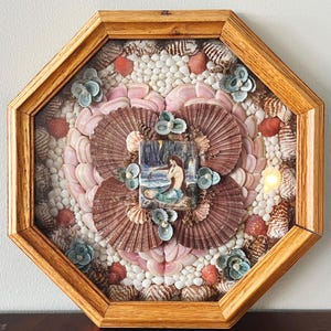 May include: An octagonal wooden framed seashell art piece. The artwork features a central image of a mermaid, surrounded by a pattern of pink, brown, and white seashells, with accents of blue and red shells.