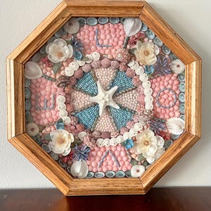 Puede incluir: Una obra de arte enmarcada octogonal con un diseño central de estrella de mar blanca, rodeada de conchas marinas azules y rosas. La obra de arte incluye flores blancas y está sobre un fondo rosa. El marco es de madera marrón claro.
