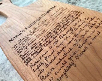 May include: A wooden cutting board with a handwritten recipe for "Nana's Famous Cookies" engraved on the surface. The recipe includes ingredients like butter, sugar, almond extract, and flour. The board has a handle and is made of light wood.
