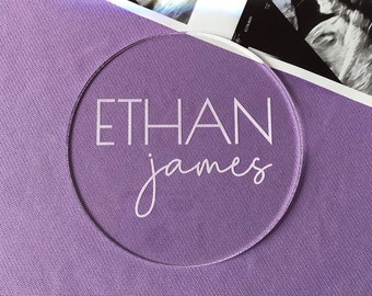 Op de afbeelding: Een ronde, transparante acrylplaat met de naam "ETHAN james" in wit.