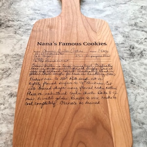 Op de afbeelding: Een houten snijplank met een handgeschreven recept voor "Nanas Beroemde Koekjes" erop. Het recept bevat ingredi&euml;nten zoals boter, suiker, amandel extract en bloem. Het recept bevat ook instructies voor het cremen van de boter en suiker, het koelen van het deeg, het uitrollen en het bakken van de koekjes.