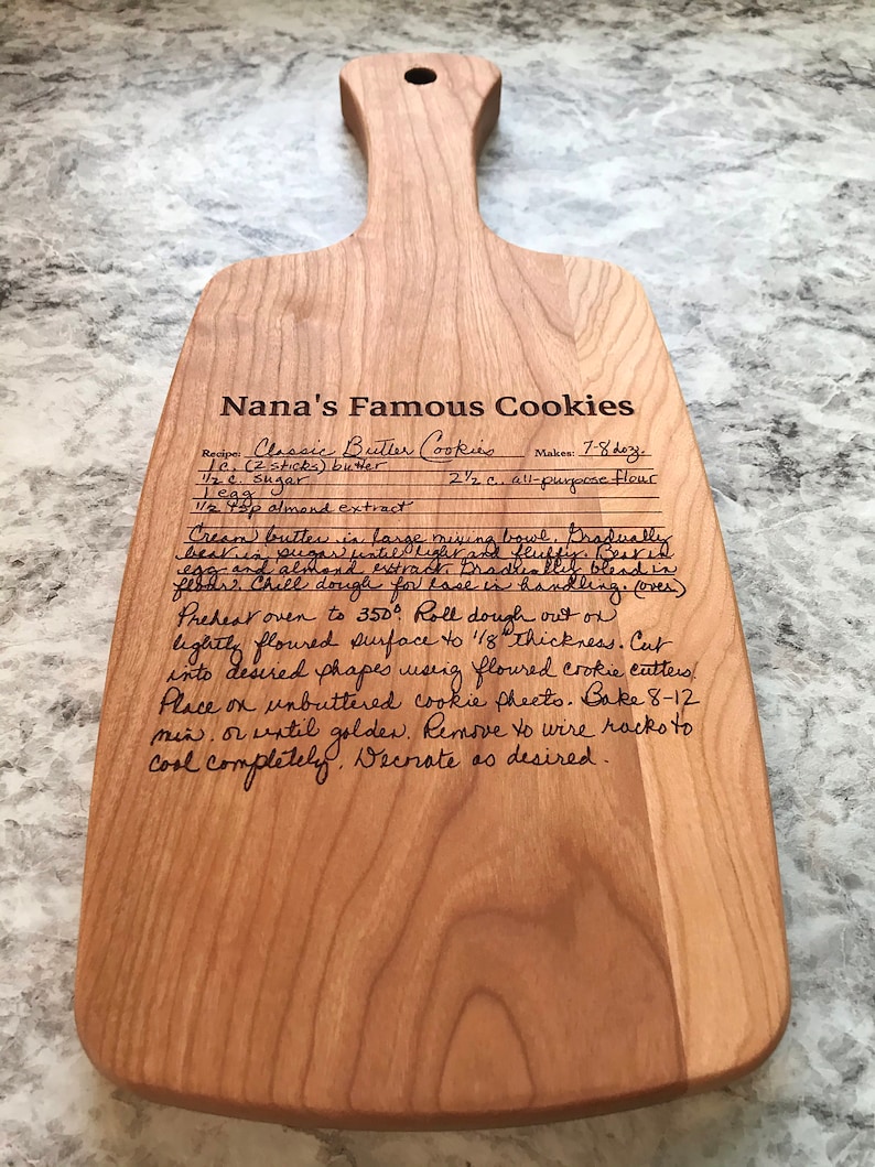 Op de afbeelding: Een houten snijplank met een handgeschreven recept voor "Nanas Beroemde Koekjes" erop. Het recept bevat ingredi&euml;nten zoals boter, suiker, amandel extract en bloem. Het recept bevat ook instructies voor het cremen van de boter en suiker, het koelen van het deeg, het uitrollen en het bakken van de koekjes.