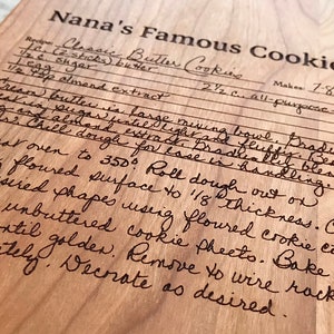 Op de afbeelding: Een houten snijplank met een handgeschreven recept voor "Nanas Beroemde Koekjes" gegraveerd op het oppervlak. Het recept bevat ingredi&euml;nten zoals boter, suiker, amandel extract en bloem. Het bevat ook instructies voor het mengen, koelen, uitrollen, uitsteken, bakken en decoreren van de koekjes.