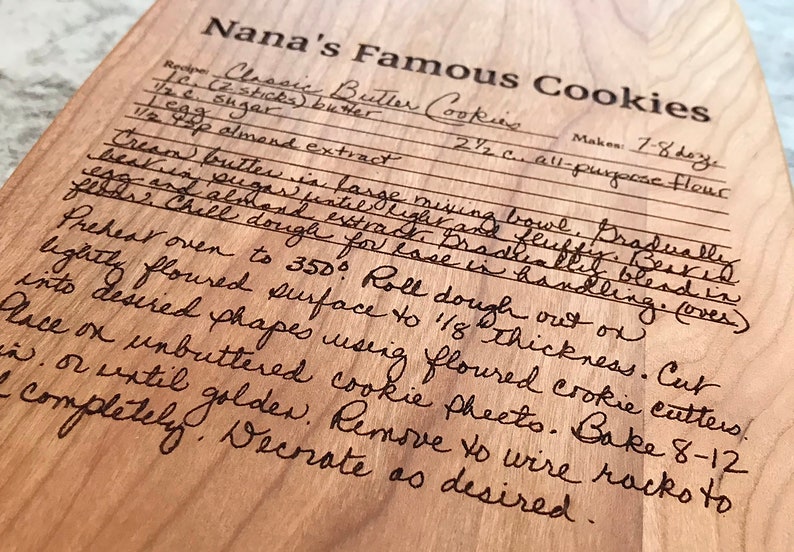Op de afbeelding: Een houten snijplank met een handgeschreven recept voor "Nanas Beroemde Koekjes" gegraveerd op het oppervlak. Het recept bevat ingredi&euml;nten zoals boter, suiker, amandel extract en bloem. Het bevat ook instructies voor het mengen, koelen, uitrollen, uitsteken, bakken en decoreren van de koekjes.