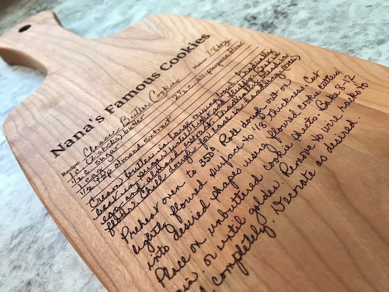 Op de afbeelding: Een houten snijplank met een handgeschreven recept voor "Nanas Beroemde Koekjes" gegraveerd op het oppervlak. Het recept bevat ingredi&euml;nten zoals boter, suiker, amandel extract en bloem. De plank heeft een handvat en is gemaakt van licht hout.