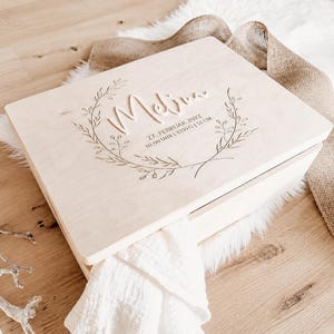 Könnte beinhalten: Eine hellfarbene Holz-Andenkenbox mit dem Namen "Melina" und Geburtsdaten auf dem Deckel graviert. Der Deckel hat ein floralen Kranz-Design. Die Box liegt auf einer weißen, fellartigen Oberfläche.