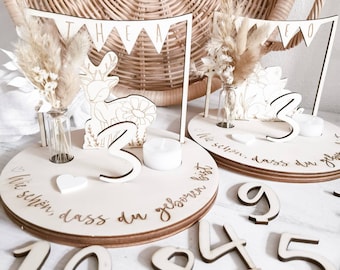 May include: Wooden birthday decorations with a deer, flowers, and a banner that says "THEA". The decorations are on a wooden base with the words "Wie schön, dass du geboren bist" and the numbers 1, 2, 8, 4, and 5.