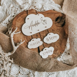 Könnte beinhalten: Ein hölzernes Schild in Wolkenform mit dem Namen "Aedda" und einer Lama-Illustration. Das Schild hängt an einem runden Holzstück mit drei weiteren wolkenförmigen Schildern mit dem Text "11.01.24", "51cm" und "2760g".