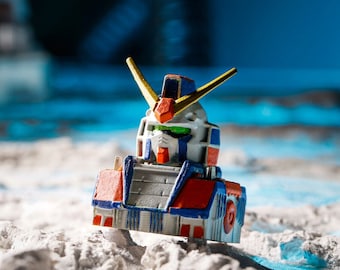 May include: A white, blue, and red robot toy with gold accents. The robot has a large head with a green visor and is standing on a white, sandy surface.