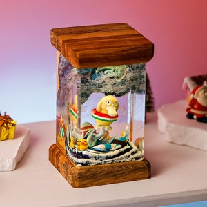 May include: A decorative snow globe with a wooden base and top. Inside, a yellow duck figure with a red and green life preserver is surrounded by a miniature ocean scene. The globe is set on a white shelf with Christmas-themed decorations.
