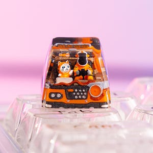 Könnte beinhalten: Eine individuelle orangefarbene und schwarze Tastenkappe mit einer Miniaturszene von zwei Figuren in einem Fahrzeug. Die Tastenkappe hat eine klare, transparente Oberseite und ist auf einer transparenten Tastatur angebracht. Die Figuren sind eine Katze und eine Person in einem orangefarbenen Anzug.