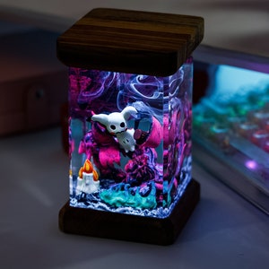 May include: A decorative lamp with a wooden base and top, featuring a clear, rectangular body. Inside, a miniature scene with a white cartoon character, pink and purple elements, and a small candle is illuminated with colorful lights.