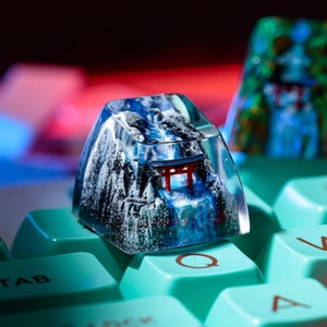 May include: A custom keycap featuring a mountain landscape with a waterfall and a red torii gate. The keycap is clear with a blue and white design, sitting on a light blue keyboard. Another keycap with a similar design is visible in the background.
