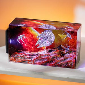 Peut inclure: Un bloc de résine rectangulaire représentant une scène de Star Wars avec le Faucon Millenium et des chasseurs X-wing sur fond de planète rouge et orange. Le bloc est posé sur une surface blanche.