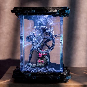 May include: A rectangular, illuminated display case featuring a Predator and an Alien in a battle scene. The figures are detailed and dark, set against a blue, smoky background. The case has a black, textured base and top.