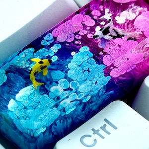 May include: A custom keycap with a koi carp design. The keycap has a layered resin design with a yellow and black koi carp swimming in a blue and purple ocean scene. The keycap is set against a white keyboard.