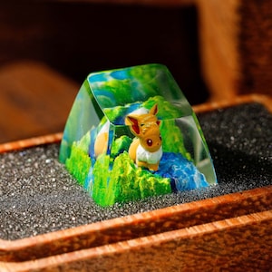 May include: A custom artisan keycap featuring a miniature Eevee figure inside a clear, geometric resin enclosure. The keycap has a landscape design with green, blue, and yellow colors, and is set in a wooden box.