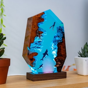 Könnte beinhalten: Eine Holz- und Harzlampe mit einer blauen und weißen Ozeanszene. Die Szene zeigt einen Taucher und drei Haie. Die Lampe hat einen Holzsockel.